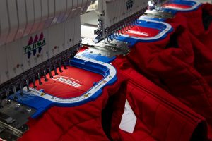 The embroidery machinery at Tacom's best professional custom corporate wear company, stitching high-quality embroidered logos onto red shirts.